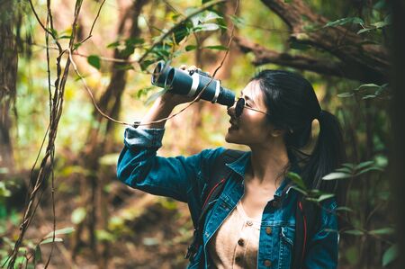 Beautiful Asian woman with binoculars telescope in forest looking destination. People lifestyles and leisure activity. Nature and backpacker traveling jungle background. Bird watching and livingの写真素材