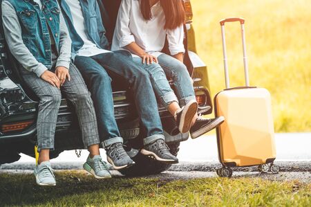 Closeup lower body of group of friends relaxing on SUV car trunk with trolly luggage along road trip with autumn mountain hill background. Freedom road way. People lifestyle transportation travelの写真素材