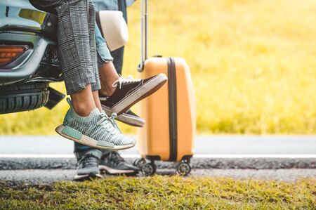 Closeup lower body of group of friends relaxing on SUV car trunk with yellow trolly luggage along road trip with mountain hill background. Freedom road way. People lifestyle and transportation travelの写真素材