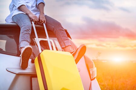 Closeup of happy Asian woman on top of car with luggage bag. Girl sitting on roof and looking sunset before evening. People lifestyle in long vacation trip concept. Nature and transportation vehicleの写真素材