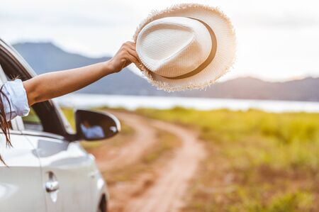 Happy woman hand holding hat outside open window car with meadow and mountain lake background. People lifestyle relaxing as traveler on road trip in holiday vacation. Transportation and travel conceptの写真素材