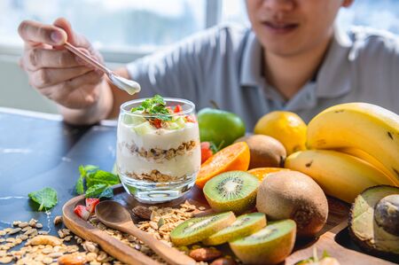 Close up of many fruit yogurt with granola and man scooping hand on table in kitchen background. Food and beverage ingredient concept. Happy vegetarian man hungry and ready to eat vegetable vitamin cの写真素材