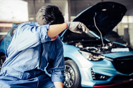 Car mechanic resting from exhaustion and wiping sweat from tired hard work in auto repair shop garage. Business industry and transportation concept. People occupation and job. Automobile technicianの写真素材