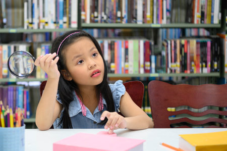 Asian girl holding magnifying glass and thinking in library at school. Education and learning conceptの写真素材