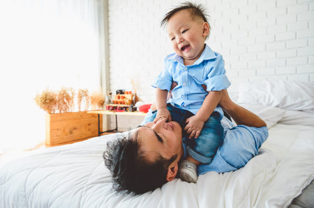 Two Asian father and his son are lying and playing in bed at bedroom. Child on top of Japanese dad body happily. Leisure time. People lifestyle health affection love.の写真素材