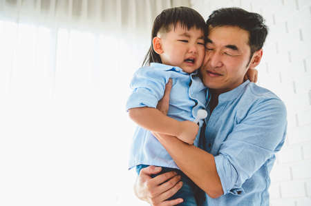 Asian Japanese father carrying and consoling his crying son in bedroom at his home with window and white curtain background. People lifestyle health. Quarantine in Covid-19 or Coranavirus epidemicの写真素材