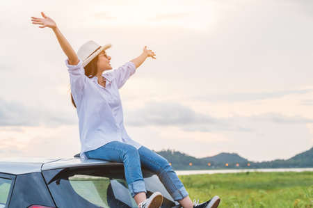 Happy Asian woman spread arms on car roof under sunset at seaside with nature  background during travel in holiday. Confidence cheerful girl with dawn sky. Sun sea and mountain. Vacation conceptの写真素材
