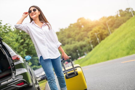 Beauty Asian woman with sunglasses dragging yellow suitcase baggage alone on road trip. People lifestyle and vacation concept. Nature and summer background. Girl having car adventure transportationの写真素材