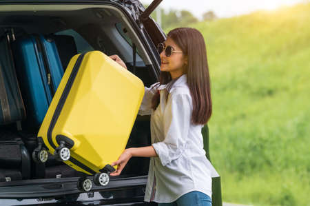 Asian woman lifting yellow suitcase into SUV car during travel in long weekend trip. People lifestyles and transportation concept. Girl put luggage from car trunk to camping. Selective focus on carの写真素材