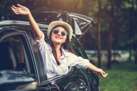 Happy woman waving hand outside open window car with meadow and mountain forest background. People lifestyle relaxing as traveler on road trip in holiday vacation. Transportation and weekend travelの写真素材