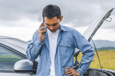 Asian man calling car maintenance service after having accident during travel long weekend between road trip. Car broken in mountain meadow background. People transportation. Need help insurance claimの写真素材