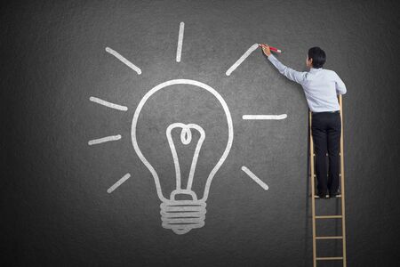 businessman standing on a ladder drawing light bulbの写真素材