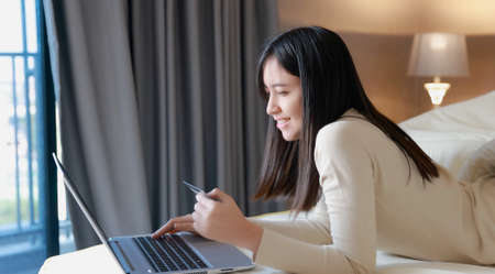 Asian woman use laptop on bed at home for online shopping with credit cardの写真素材