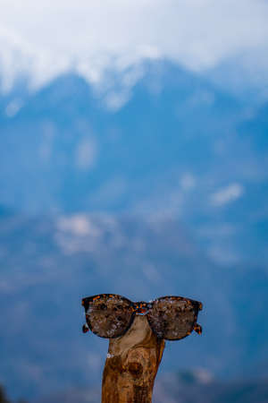 A Sun Glasses is placed on a wood in front of the mighty mountain with selective focus.
