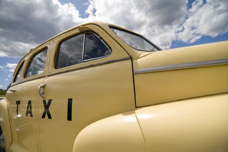 Side view of a pale yellow, retro styled classic taxi cab. Wide angle view with light fluffy clouds against a clear blue sky.の写真素材