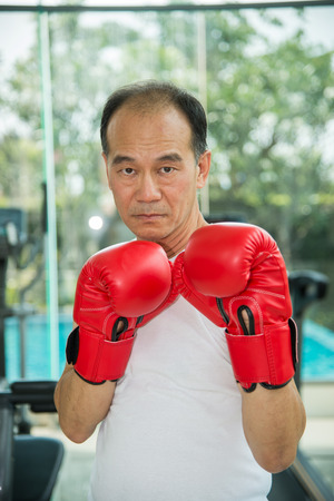 Old man wearing red boxing gloves exercising in fitness or gym, look to camera ready to fightingの写真素材