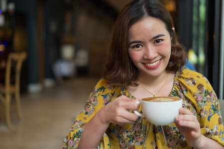 Portrait of beautiful woman and holding a cup of coffee in her hand in blur background coffee shop, she drink coffee in the morningの写真素材