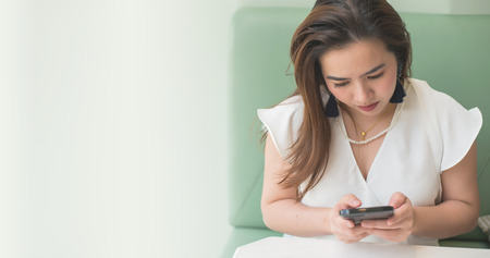 beautiful Asian woman wear white shirt on green sofa using mobile phone chat with friend and blurred background with copy space for textの写真素材