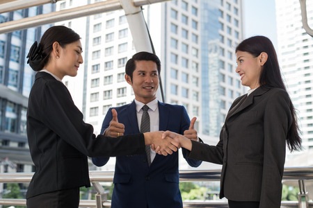 business man show thumb up and two business woman shaking hands for demonstrating their agreement to sign agreement or contract between their firms, companies, enterprises. success conceptの写真素材