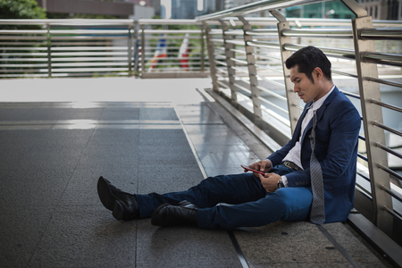businessman unemployed from company sitting on street and using mobile phone, concept of business failure and unemployment problemの写真素材