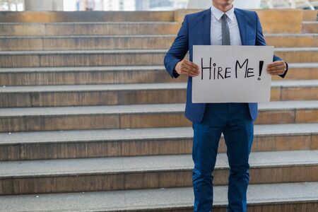 Hire me! Handsome businessman wear blue suit holding poster with hire me text message while standing outdoors and against building structureの写真素材