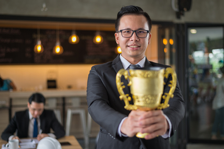 Asian businessman raising up hand and holding a golden trophy cup to cheerful and celebrated his successful in career and mission. concept of winner and competition.の写真素材