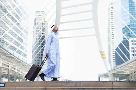 Muslim man wear white turban and dress walking with luggage in public place and modern building background. concept of business travel.の写真素材