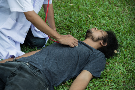 doctor using stethoscope to listen heart beat of a man, who heart attack on green grass in the garden.の写真素材
