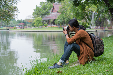 male travelers sitting on green grass near pond in public park, he holding camera on hand and take a photo. concept of travel, tripの写真素材