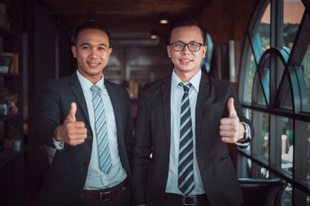 Two Asian businessman stand and smile and show thumb up at the office, successful conceptの写真素材