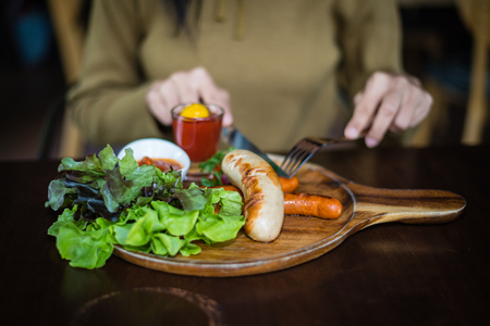 Blurred hand of Woman holding knife and fork for eating food, close up Sausage with Tomato Sauce and green vegetable on wood plateの写真素材