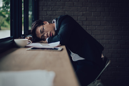 Tired businessman sleeping on counter bar at coffee shop with document, laptop and cup of coffee.の写真素材