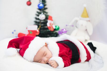 little baby wearing Santa Claus costume sleep on white fur carpet with Christmas tree. Concept of celebrates Christmas and New Year's holidays.の写真素材