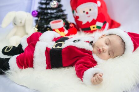 little baby wearing Santa Claus costume sleep on white fur carpet with Christmas tree. Concept of celebrates Christmas and New Year's holidays.の写真素材