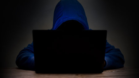 Man wears a blue hooded shirt in front of a laptop computer. Hacking ideas, computer theft to damage