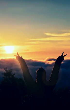 A woman raises her arms and holds two fingers behind the morning sun. Concept of struggle and commitment.の写真素材