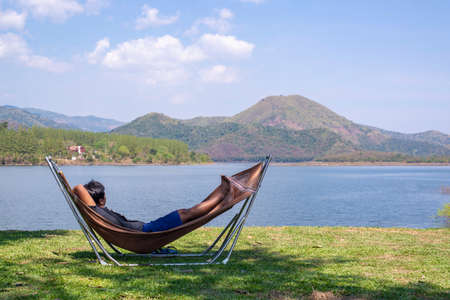 A man sleeps in a hammock on a green grass by a lake, a mountain view and a bright sky. Relaxation ideas, life planning for future comfort.の写真素材