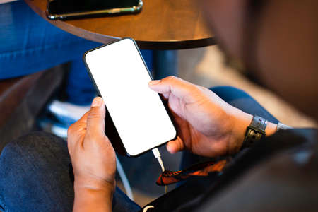 A blank white smartphone screen is charging the battery in the hand of an Asian man at a table in a coffee shop. The use of social media, internet networkの写真素材