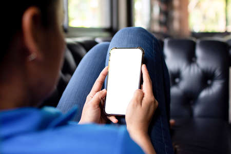 A young Asian woman sitting on a smartphone with a blank white screen for text input and design on the sofa at home.の写真素材