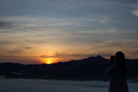 A dark silhouette of a tourist using a camera to capture the light of the morning sun.の写真素材
