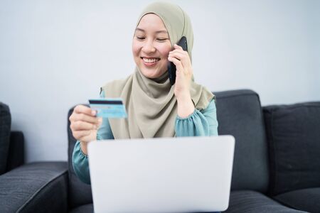 Beautiful muslim woman talking on phone while doing online transaction on her laptop.