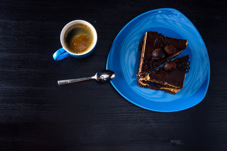 A piece of chocolate cake on a blue plate. Near a cup of coffee. All on a black wooden background.