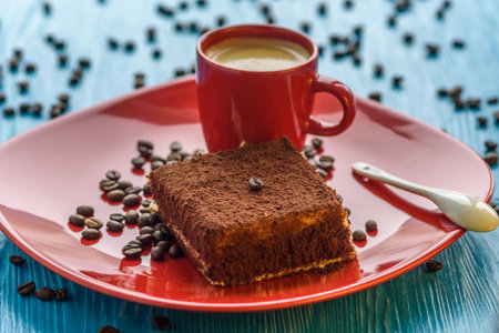 Tiramisu cake on a red plate, near a cup of coffee, photographed on a light wooden background.の写真素材