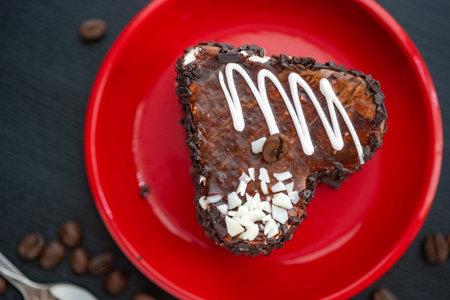 A delicious heart-shaped cake sprinkled with pieces of chocolate lies on a red plate. Photographed close-up on a wooden background.の写真素材