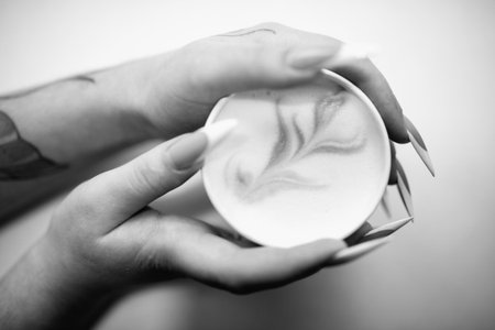 Hands holding a white cup of latte art with intricate designs, showing creamy texture and warm...