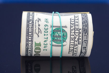 Rolled stack of hundred dollar bills secured with a rubber band, resting on a reflective surface, symbolizing wealth, finance, and economic prosperity in a minimalist settingの写真素材