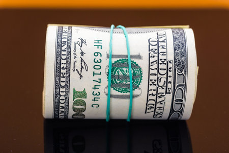 Rolled stack of hundred dollar bills secured with a rubber band, resting on a reflective surface, symbolizing wealth, finance, and economic prosperity in a minimalist settingの写真素材
