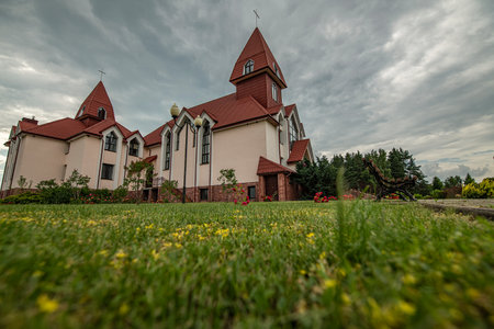 Beautiful modern church with a unique architectural design, featuring a prominent steeple, surrounded by vibrant flowers and lush greenery, under a dramatic cloudy skyの写真素材