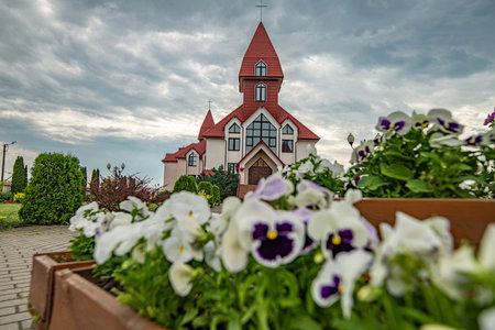 Beautiful modern church with a unique architectural design, featuring a prominent steeple, surrounded by vibrant flowers and lush greenery, under a dramatic cloudy skyの写真素材