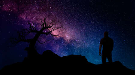 single dead tree and man silhouette on hill at night with milky way core and stars background